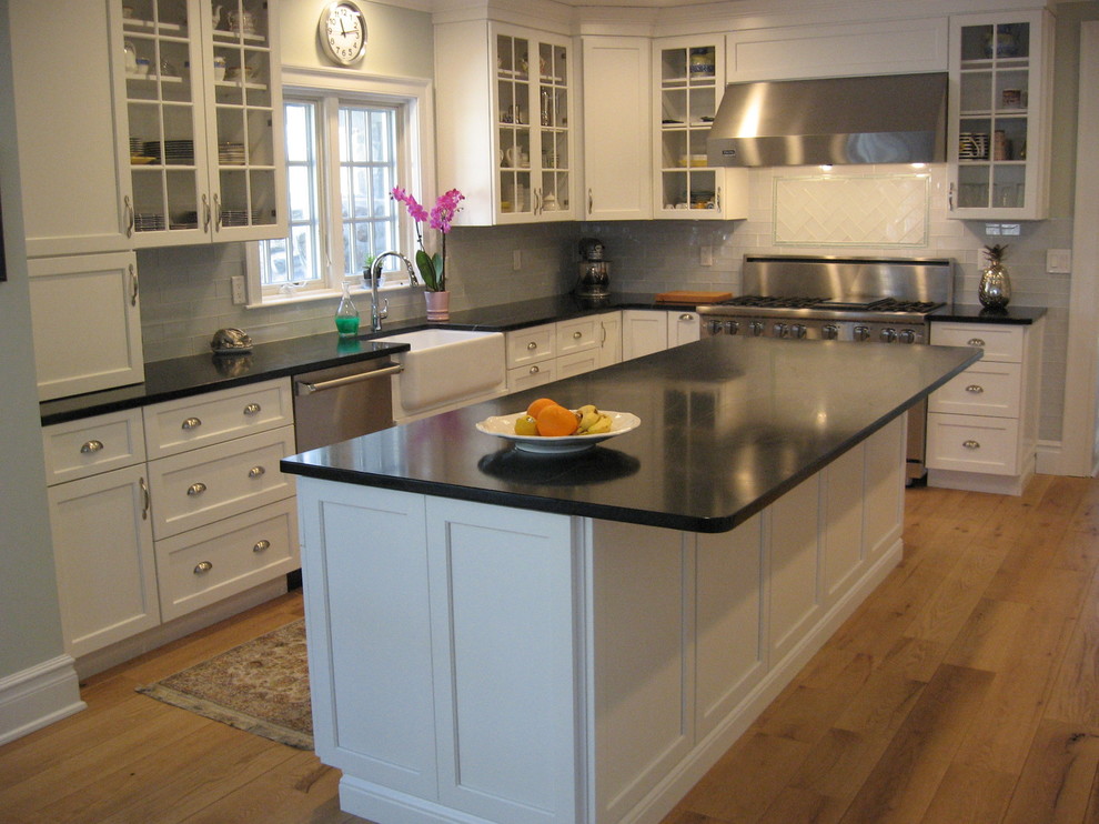 This is an example of a medium sized traditional l-shaped kitchen/diner in New York with a belfast sink, shaker cabinets, white cabinets, soapstone worktops, grey splashback, ceramic splashback, stainless steel appliances, light hardwood flooring, an island, brown floors and black worktops.