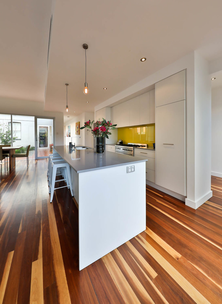 Photo of a medium sized contemporary galley open plan kitchen in Adelaide with a double-bowl sink, white cabinets, engineered stone countertops, yellow splashback, glass tiled splashback, stainless steel appliances, medium hardwood flooring, an island and flat-panel cabinets.