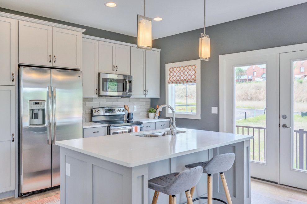 Photo of a classic galley kitchen in DC Metro with a double-bowl sink, shaker cabinets, white cabinets, beige splashback, stainless steel appliances, light hardwood flooring and an island.