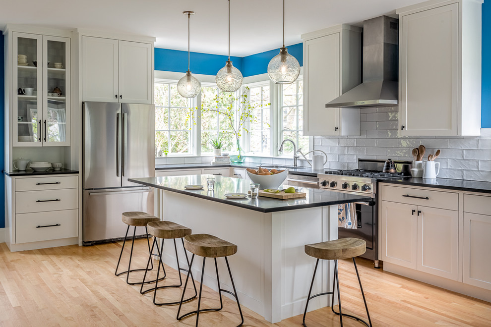Transitional l-shaped light wood floor and beige floor kitchen photo in Portland Maine with shaker cabinets, gray cabinets, white backsplash, subway tile backsplash, stainless steel appliances, an island and black countertops