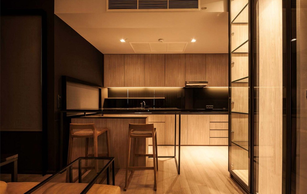 Example of a mid-sized minimalist single-wall light wood floor open concept kitchen design in New York with an undermount sink, flat-panel cabinets, light wood cabinets, solid surface countertops, black backsplash, glass sheet backsplash, paneled appliances and a peninsula