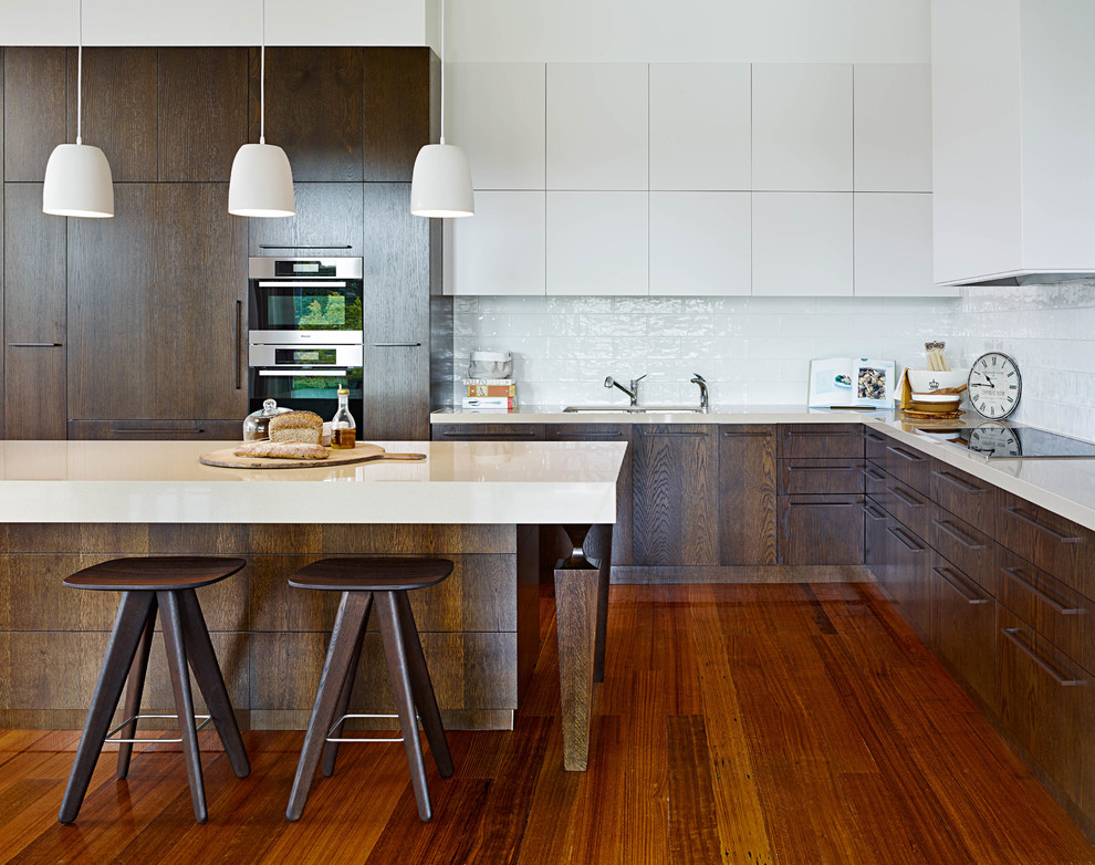 Moderne Küche in L-Form mit Doppelwaschbecken, flächenbündigen Schrankfronten, weißen Schränken, Küchenrückwand in Weiß, Küchengeräten aus Edelstahl, braunem Holzboden, Kücheninsel, Mineralwerkstoff-Arbeitsplatte und Rückwand aus Porzellanfliesen in Melbourne