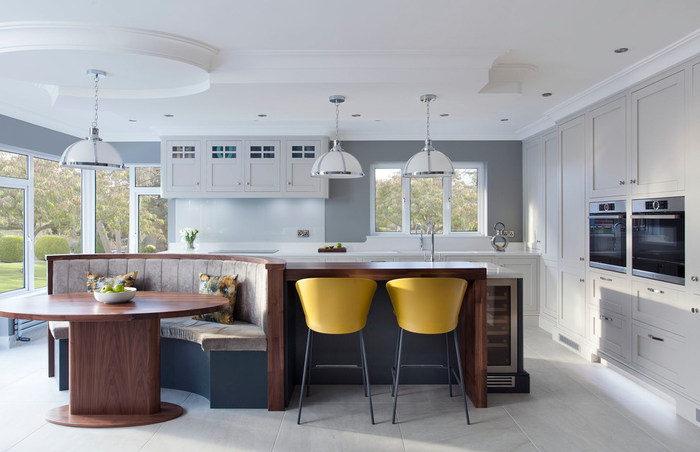 Идея дизайна: кухня в стиле неоклассика (современная классика) с обеденным столом, фасадами в стиле шейкер, серыми фасадами, белым фартуком, фартуком из стекла, техникой из нержавеющей стали и серым полом