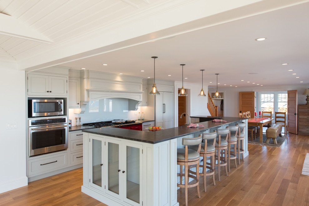 This is an example of a large coastal kitchen/diner in Boston with a belfast sink, shaker cabinets, grey cabinets, granite worktops, white splashback, glass sheet splashback, stainless steel appliances, medium hardwood flooring, an island and black worktops.