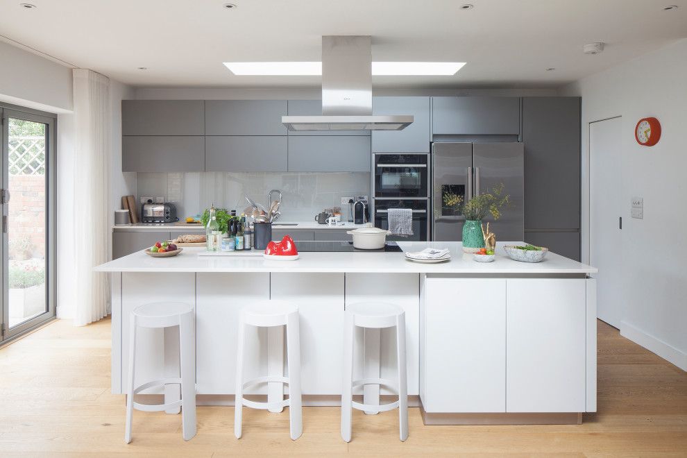 Large scandinavian single-wall open plan kitchen in Sussex with an integrated sink, flat-panel cabinets, grey cabinets, composite countertops, green splashback, glass sheet splashback, stainless steel appliances, medium hardwood flooring, an island, brown floors and white worktops.