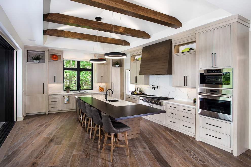 Inredning av ett modernt kök, med en undermonterad diskho, släta luckor, skåp i ljust trä, vitt stänkskydd, rostfria vitvaror, mörkt trägolv, en köksö och brunt golv