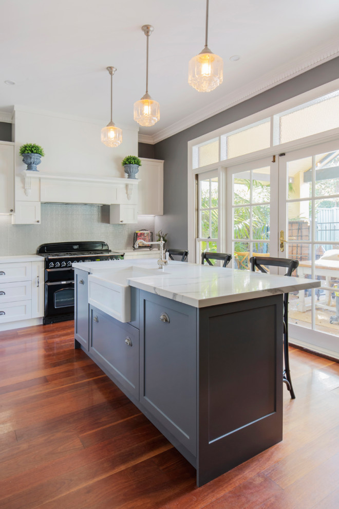 This is an example of a medium sized classic l-shaped kitchen/diner in Sydney with a belfast sink, shaker cabinets, white cabinets, engineered stone countertops, grey splashback, metal splashback, black appliances, medium hardwood flooring, an island, brown floors and multicoloured worktops.