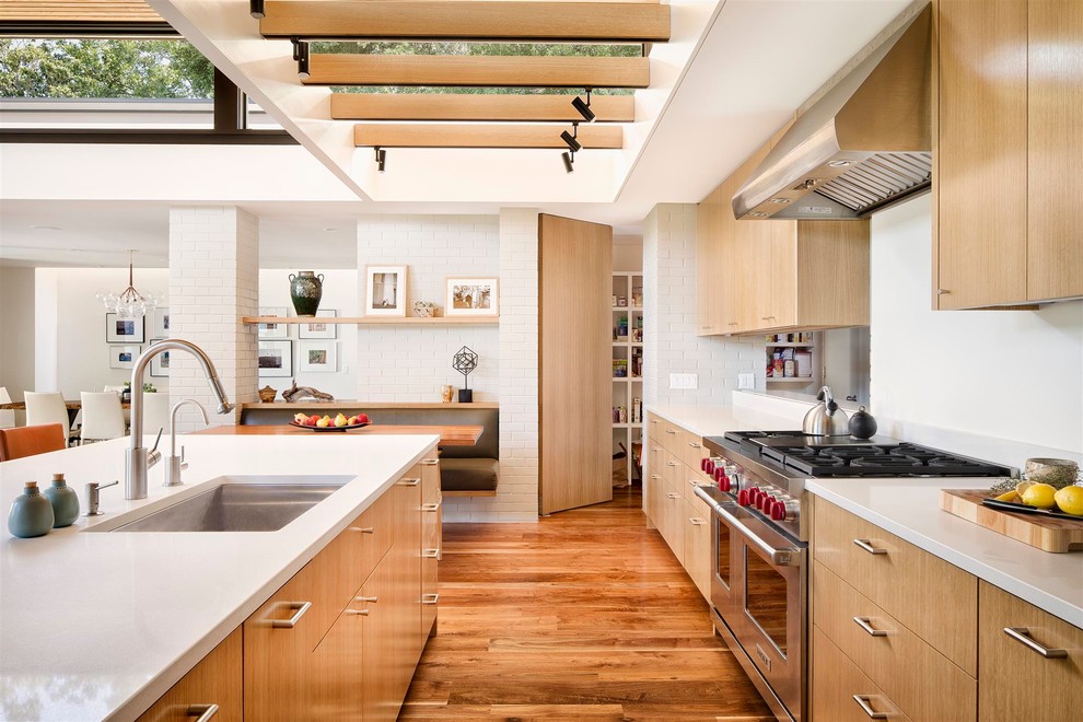 Cette image montre une cuisine design en bois clair avec un évier encastré, un placard à porte plane, un électroménager en acier inoxydable, un sol en bois brun et îlot.