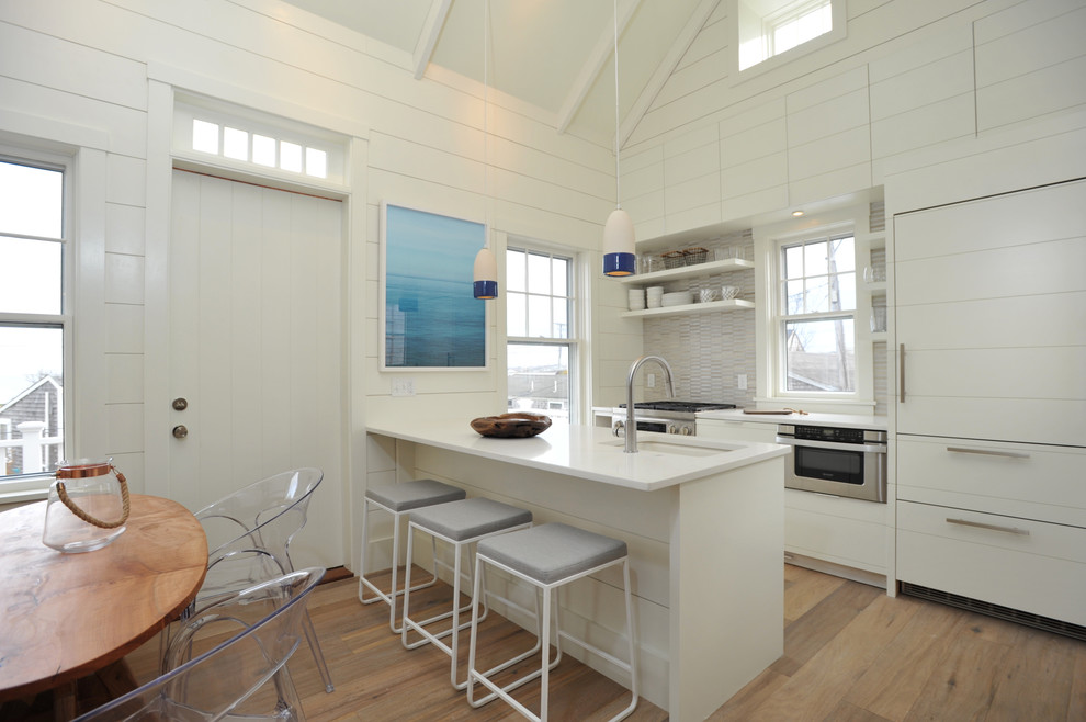 Foto på ett maritimt kök och matrum, med en undermonterad diskho, vita skåp, grått stänkskydd, rostfria vitvaror, ljust trägolv, en halv köksö och beiget golv