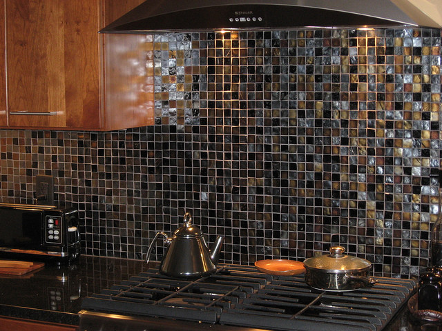 Mittelgroße Moderne Küche mit flächenbündigen Schrankfronten, hellbraunen Holzschränken, Küchenrückwand in Schwarz, Rückwand aus Glasfliesen und schwarzer Arbeitsplatte in Grand Rapids