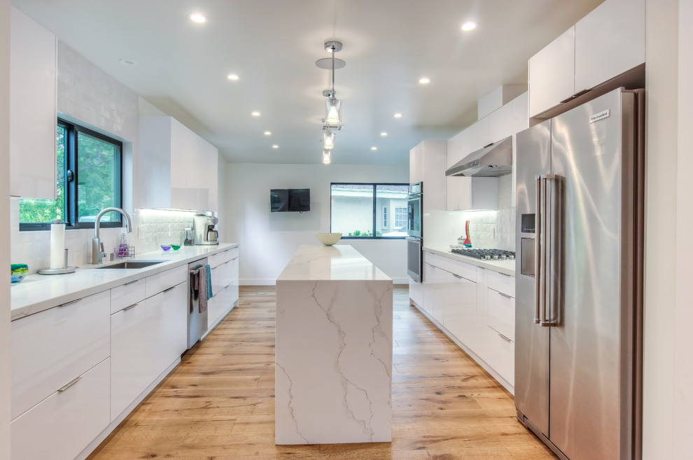 Design ideas for a contemporary kitchen in Los Angeles with flat-panel cabinets, white cabinets, engineered stone countertops, white splashback, porcelain splashback, stainless steel appliances, light hardwood flooring, an island, beige floors and white worktops.