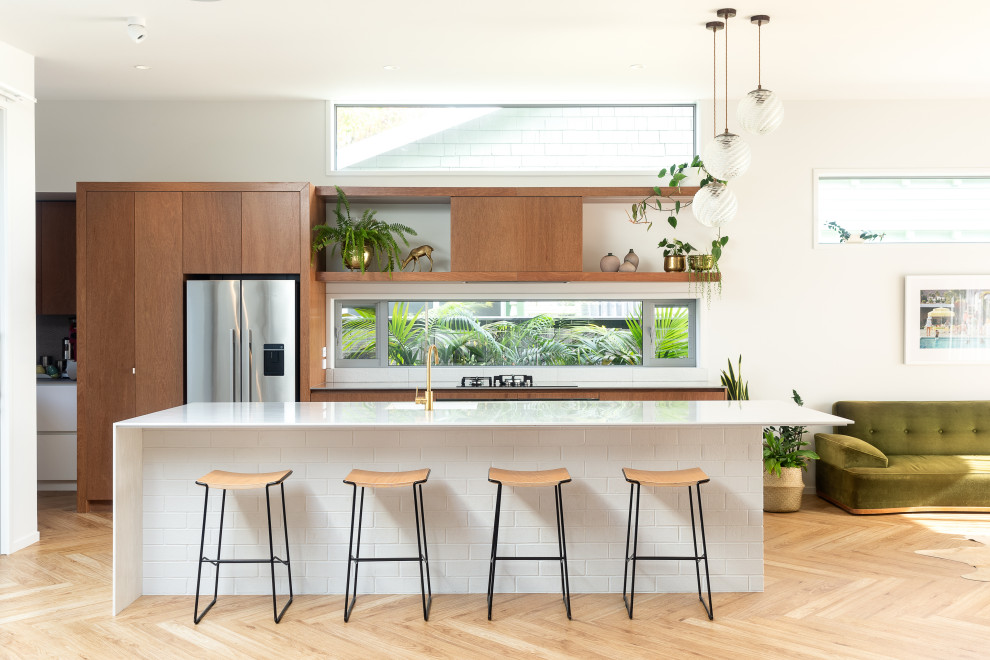 Design ideas for a contemporary galley open plan kitchen in Auckland with a submerged sink, flat-panel cabinets, medium wood cabinets, window splashback, stainless steel appliances, light hardwood flooring, an island, beige floors and white worktops.