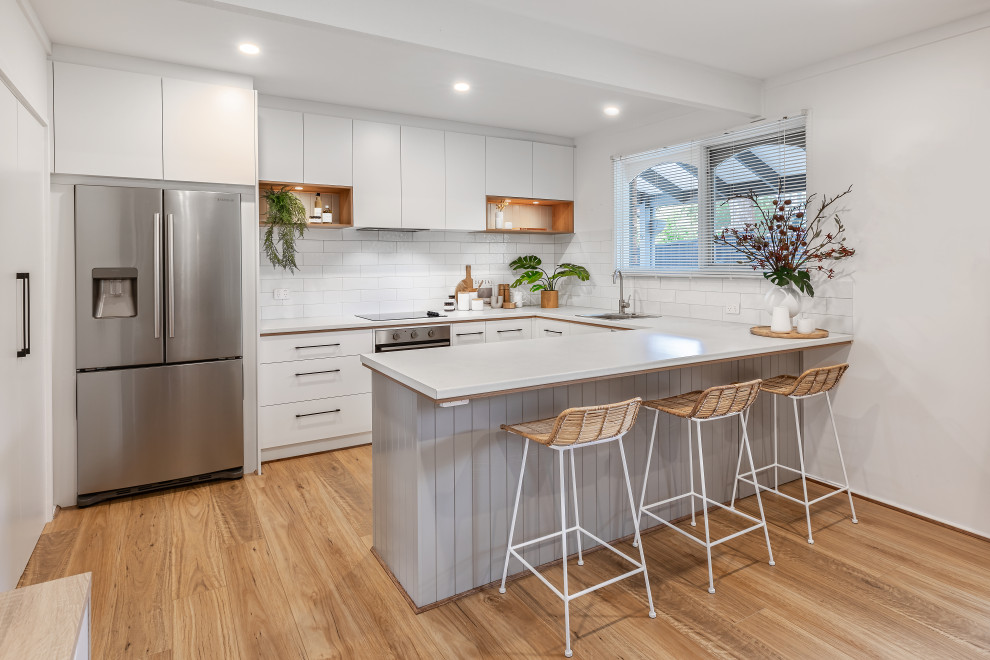 Photo of a medium sized contemporary u-shaped kitchen/diner in Adelaide with a single-bowl sink, white cabinets, laminate countertops, white splashback, metro tiled splashback, stainless steel appliances, medium hardwood flooring, a breakfast bar, brown floors, white worktops and flat-panel cabinets.