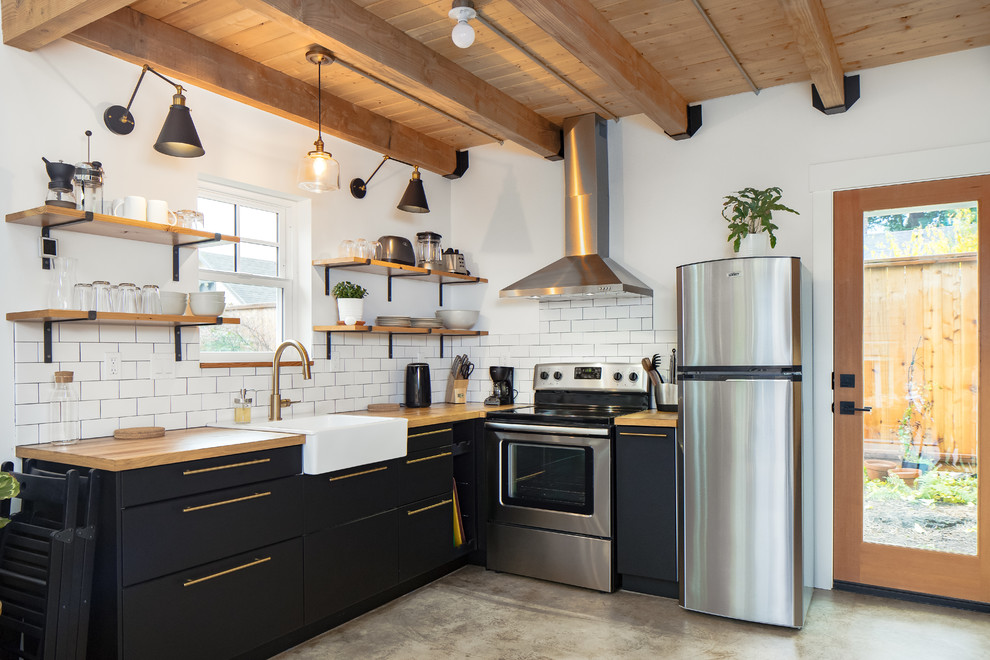 Bild på ett lantligt l-kök, med en rustik diskho, släta luckor, svarta skåp, träbänkskiva, vitt stänkskydd, stänkskydd i tunnelbanekakel, rostfria vitvaror, betonggolv och grått golv