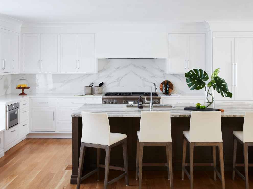 Inspiration for a large traditional l-shaped open plan kitchen in New York with a submerged sink, shaker cabinets, white cabinets, white splashback, porcelain splashback, stainless steel appliances, medium hardwood flooring, an island, beige floors and multicoloured worktops.