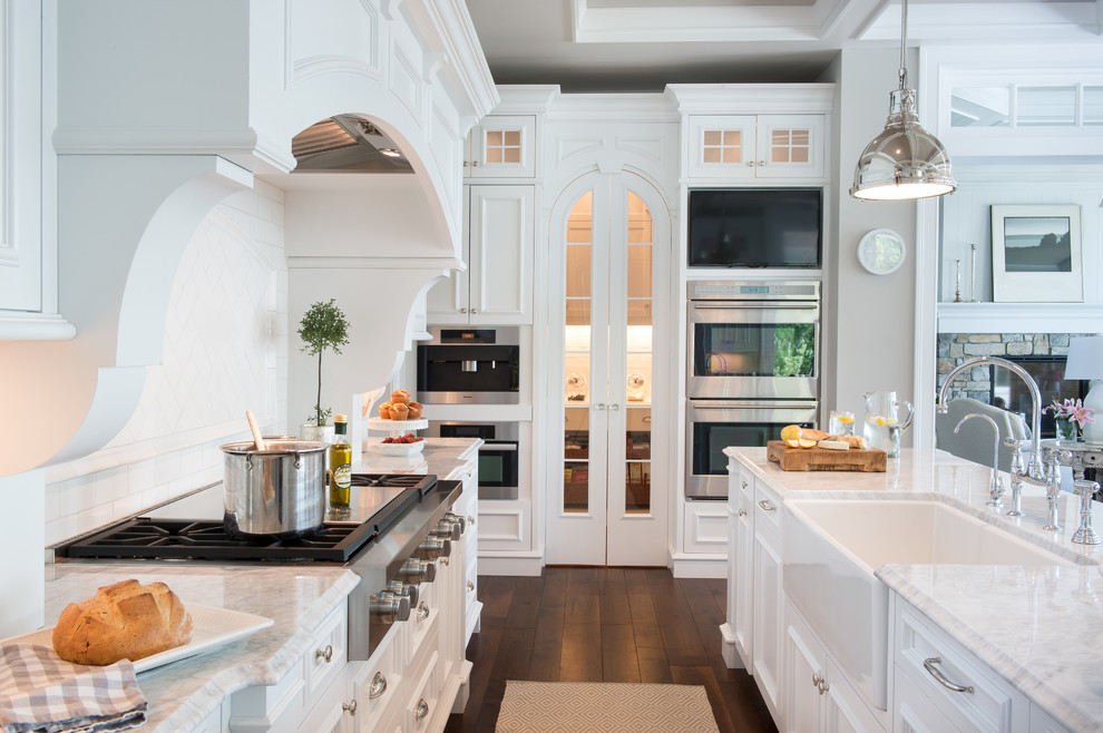 This is an example of an expansive beach style u-shaped kitchen/diner in Minneapolis with a belfast sink, shaker cabinets, white cabinets, marble worktops, white splashback, porcelain splashback, white appliances, medium hardwood flooring, multiple islands and brown floors.