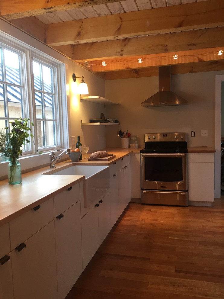 Medium sized scandinavian l-shaped enclosed kitchen in Burlington with a belfast sink, flat-panel cabinets, white cabinets, wood worktops, stainless steel appliances, light hardwood flooring and an island.