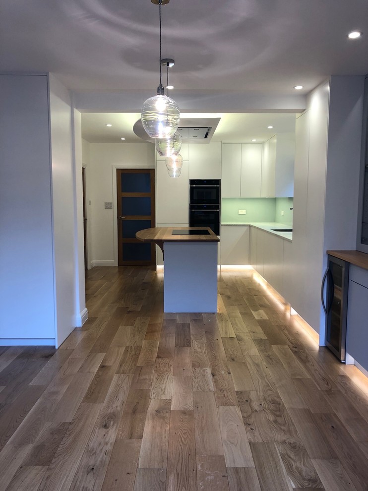 Photo of a medium sized contemporary l-shaped kitchen/diner in Hertfordshire with a built-in sink, flat-panel cabinets, white cabinets, wood worktops, grey splashback, glass tiled splashback, integrated appliances, light hardwood flooring, an island, brown floors and grey worktops.