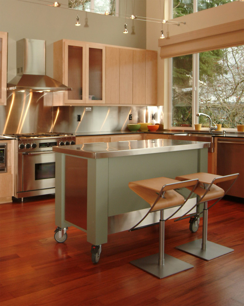 Exemple d'une cuisine américaine moderne en L et bois clair avec un évier de ferme, un placard à porte plane, une crédence métallisée, une crédence en dalle métallique, un électroménager en acier inoxydable, un sol en bois brun et îlot.