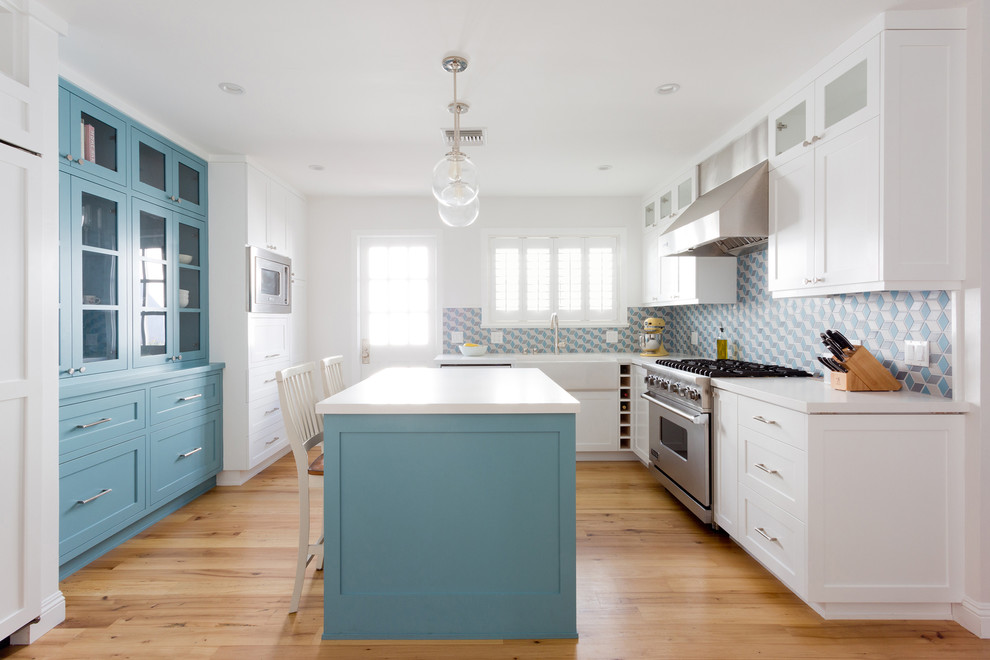 This is an example of a small classic l-shaped enclosed kitchen in Los Angeles with a belfast sink, shaker cabinets, blue cabinets, blue splashback, light hardwood flooring, an island, composite countertops, ceramic splashback, stainless steel appliances and brown floors.