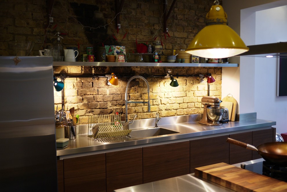Cette photo montre une cuisine américaine industrielle en bois brun de taille moyenne avec un placard à porte plane, un plan de travail en inox, une crédence en brique, un électroménager en acier inoxydable, un sol en ardoise, îlot, un sol noir et un évier intégré.