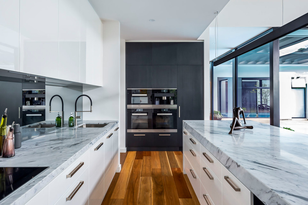 Große Moderne Küche in L-Form mit Unterbauwaschbecken, flächenbündigen Schrankfronten, weißen Schränken, Rückwand aus Spiegelfliesen, schwarzen Elektrogeräten, Kücheninsel, braunem Boden und grauer Arbeitsplatte in Adelaide