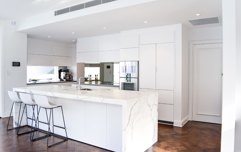 Foto på ett mycket stort funkis kök, med en undermonterad diskho, släta luckor, vita skåp, marmorbänkskiva, stänkskydd med metallisk yta, spegel som stänkskydd, rostfria vitvaror, mellanmörkt trägolv och en köksö