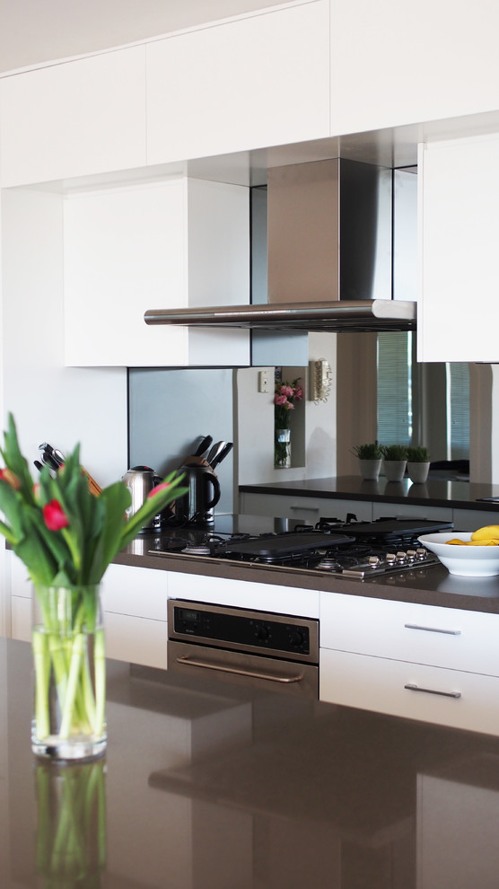 Modern inredning av ett mellanstort kök, med en undermonterad diskho, släta luckor, vita skåp, bänkskiva i kvarts, spegel som stänkskydd, rostfria vitvaror, mörkt trägolv och en köksö