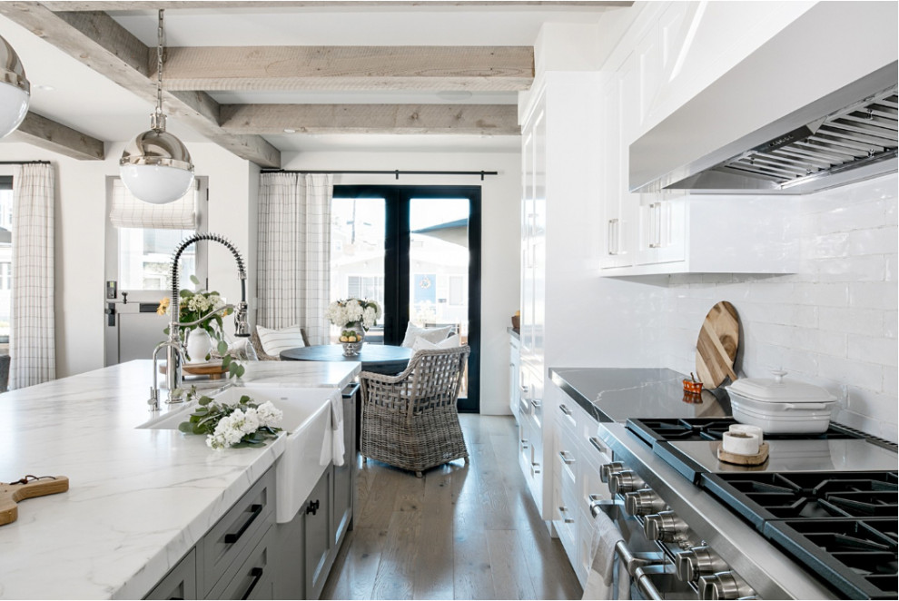 Example of a mid-sized beach style l-shaped light wood floor and beige floor open concept kitchen design in Orange County with a farmhouse sink, shaker cabinets, white cabinets, marble countertops, white backsplash, ceramic backsplash, paneled appliances, an island and white countertops