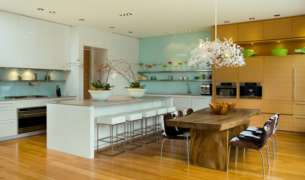 Moderne Wohnküche in L-Form mit flächenbündigen Schrankfronten, weißen Schränken, Edelstahl-Arbeitsplatte, Küchenrückwand in Blau, Glasrückwand und Küchengeräten aus Edelstahl in Toronto