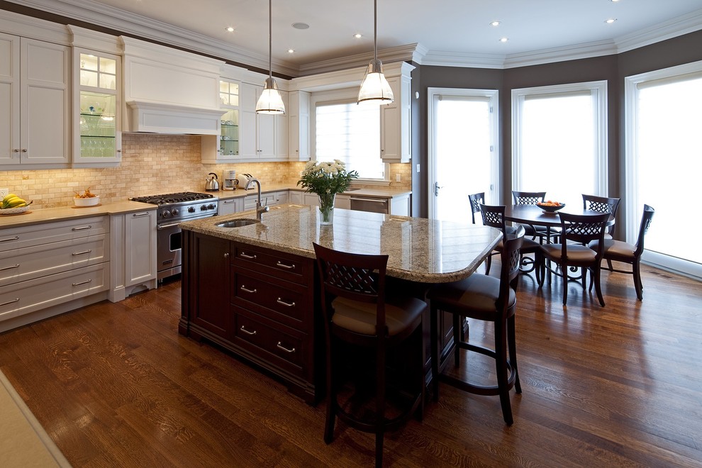 Idée de décoration pour une grande cuisine américaine tradition en U avec un placard avec porte à panneau encastré, des portes de placard blanches, un plan de travail en granite, une crédence beige, un sol en bois brun et îlot.