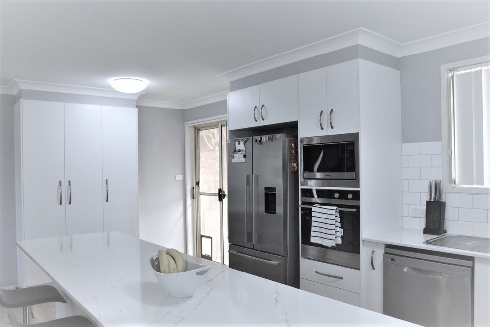 Inspiration för mellanstora moderna vitt kök, med en dubbel diskho, släta luckor, vita skåp, bänkskiva i kvarts, vitt stänkskydd, stänkskydd i tunnelbanekakel, rostfria vitvaror, mellanmörkt trägolv, en halv köksö och brunt golv