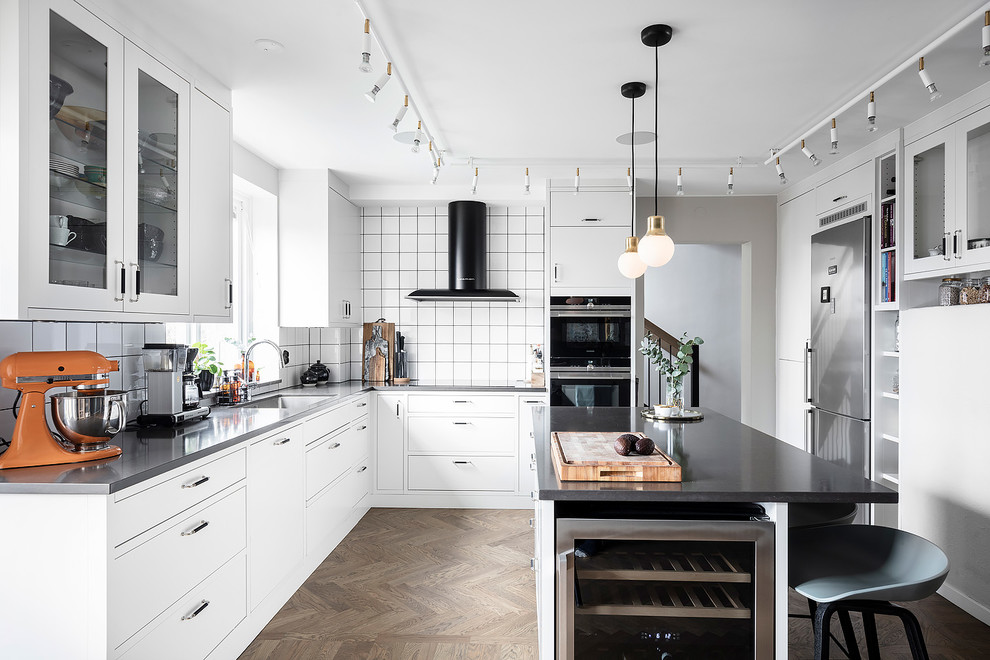 Example of a large danish u-shaped brown floor and medium tone wood floor eat-in kitchen design in Gothenburg with white cabinets, solid surface countertops, white backsplash, ceramic backsplash, stainless steel appliances, an island, black countertops and an undermount sink