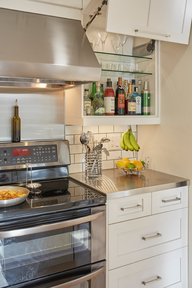Foto på ett mellanstort eklektiskt kök, med en integrerad diskho, skåp i shakerstil, vita skåp, bänkskiva i rostfritt stål, vitt stänkskydd, stänkskydd i tunnelbanekakel, rostfria vitvaror, ljust trägolv och en köksö