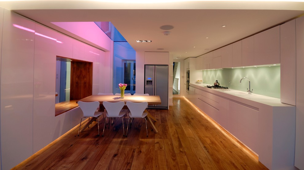 Photo of a medium sized contemporary single-wall open plan kitchen in London with an integrated sink, flat-panel cabinets, white cabinets, composite countertops, green splashback, glass sheet splashback, integrated appliances, light hardwood flooring and no island.