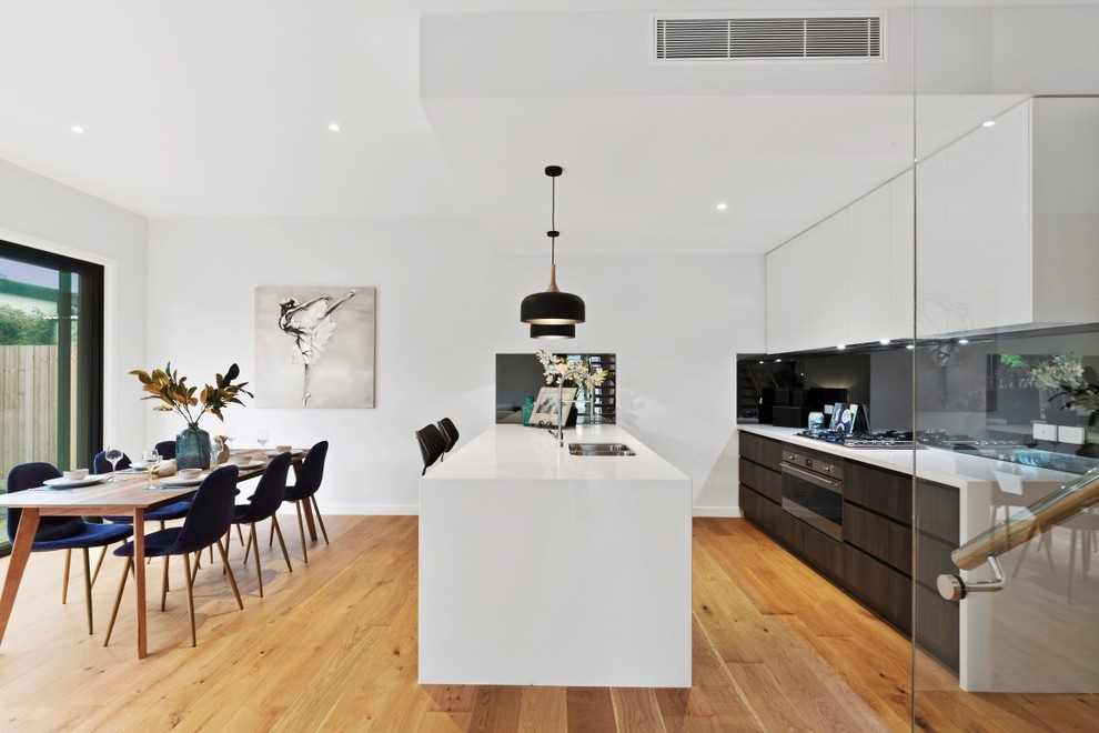 Photo of a modern galley kitchen/diner in Melbourne with a double-bowl sink, flat-panel cabinets, white cabinets, stainless steel appliances, medium hardwood flooring, an island, brown floors and white worktops.