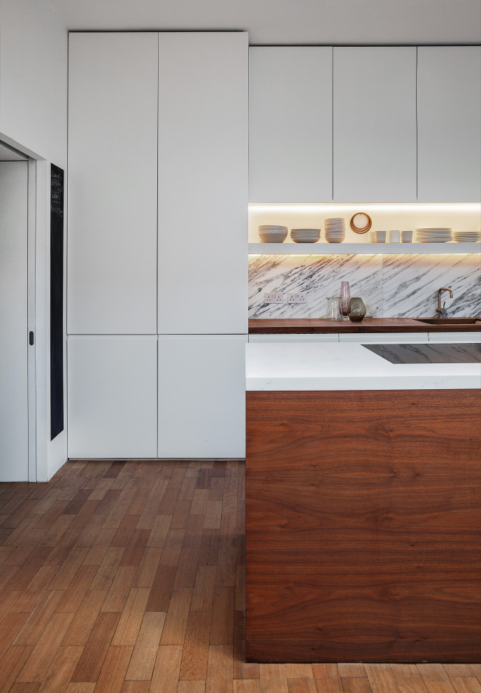 This is an example of a large contemporary single-wall open plan kitchen in London with a built-in sink, flat-panel cabinets, white cabinets, wood worktops, white splashback, marble splashback, stainless steel appliances, medium hardwood flooring, an island, brown floors and brown worktops.