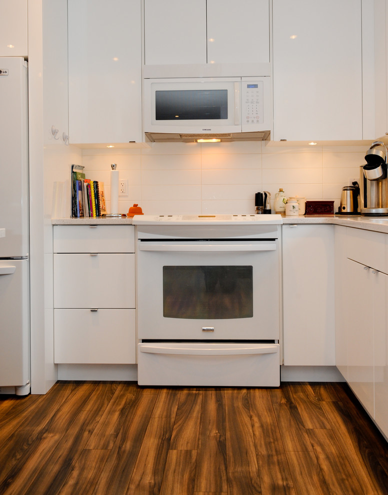 This is an example of a medium sized traditional galley kitchen/diner in Calgary with a submerged sink, flat-panel cabinets, white cabinets, engineered stone countertops, white splashback, metro tiled splashback, white appliances, medium hardwood flooring and an island.
