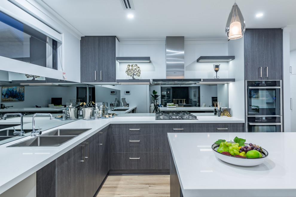 На фото: угловая кухня в современном стиле с двойной мойкой, плоскими фасадами, серыми фасадами, зеркальным фартуком, черной техникой, светлым паркетным полом, островом, бежевым полом и белой столешницей