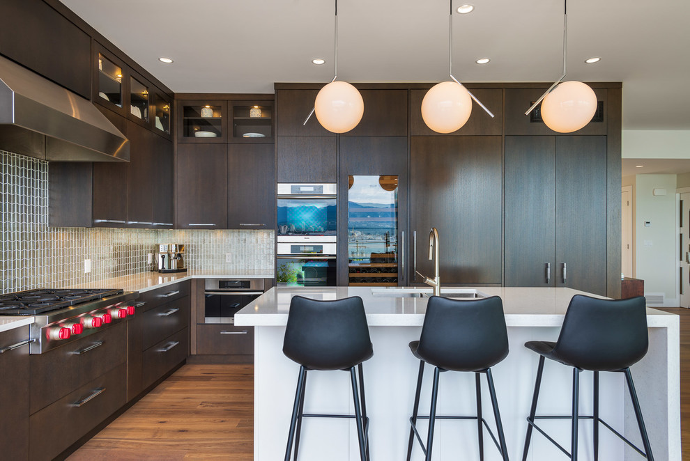This is an example of a large contemporary l-shaped kitchen/diner in Vancouver with a submerged sink, flat-panel cabinets, dark wood cabinets, quartz worktops, green splashback, glass tiled splashback, stainless steel appliances, light hardwood flooring, an island, brown floors and white worktops.