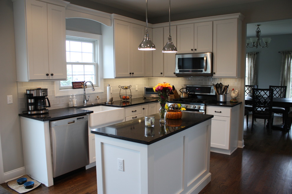 Example of a classic dark wood floor and brown floor eat-in kitchen design in New York with a farmhouse sink, recessed-panel cabinets, white cabinets, gray backsplash, glass tile backsplash, stainless steel appliances and an island