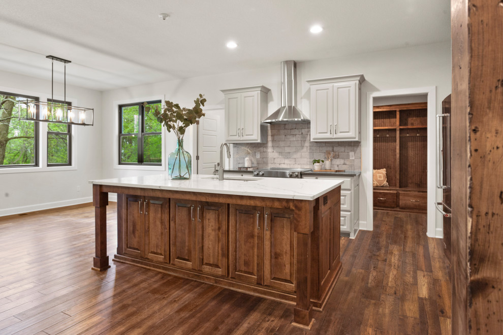 This is an example of a medium sized traditional l-shaped open plan kitchen in Kansas City with a submerged sink, raised-panel cabinets, white cabinets, engineered stone countertops, beige splashback, stone tiled splashback, stainless steel appliances, medium hardwood flooring, an island, brown floors and grey worktops.