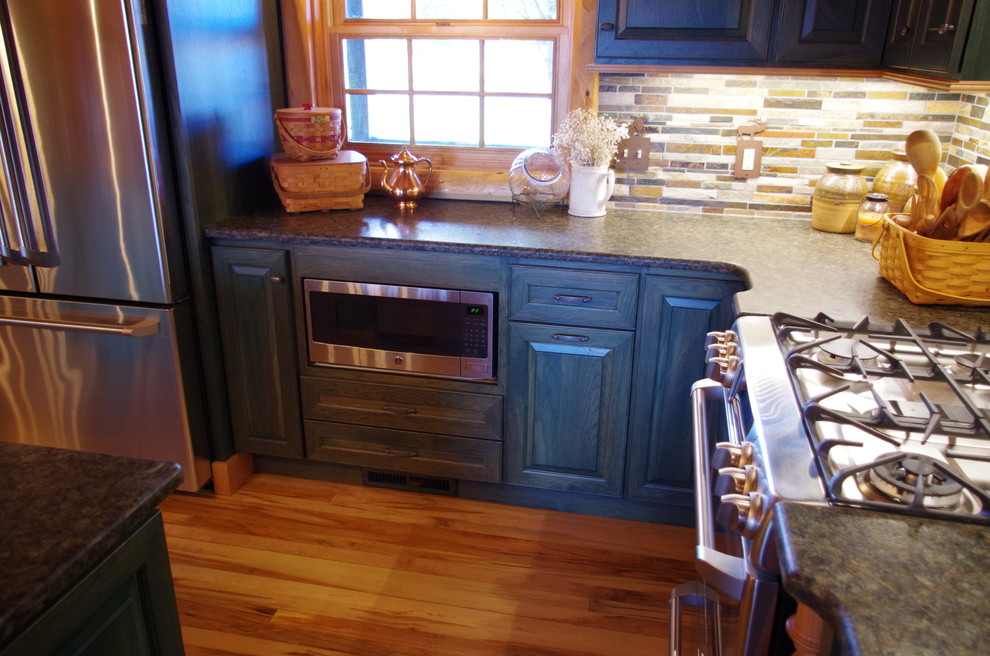 This is an example of a medium sized rustic l-shaped kitchen/diner in Other with a belfast sink, raised-panel cabinets, green cabinets, granite worktops, multi-coloured splashback, stone tiled splashback, stainless steel appliances, light hardwood flooring, an island and brown floors.
