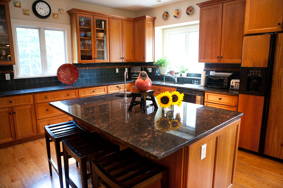 Inredning av ett klassiskt stort kök, med en köksö, luckor med infälld panel, skåp i mellenmörkt trä, granitbänkskiva, svart stänkskydd, stänkskydd i stenkakel, svarta vitvaror, en undermonterad diskho, mellanmörkt trägolv och brunt golv