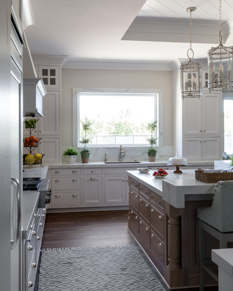 Bild på ett stort vit vitt kök, med en undermonterad diskho, luckor med infälld panel, vita skåp, marmorbänkskiva, vitt stänkskydd, stänkskydd i sten, en köksö, rostfria vitvaror, mörkt trägolv och brunt golv