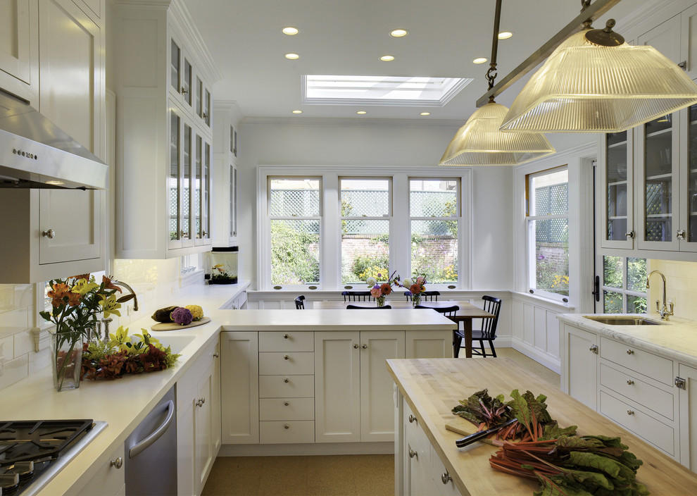 Elegant eat-in kitchen photo in San Francisco with glass-front cabinets, stainless steel appliances, solid surface countertops, white cabinets, white backsplash and subway tile backsplash