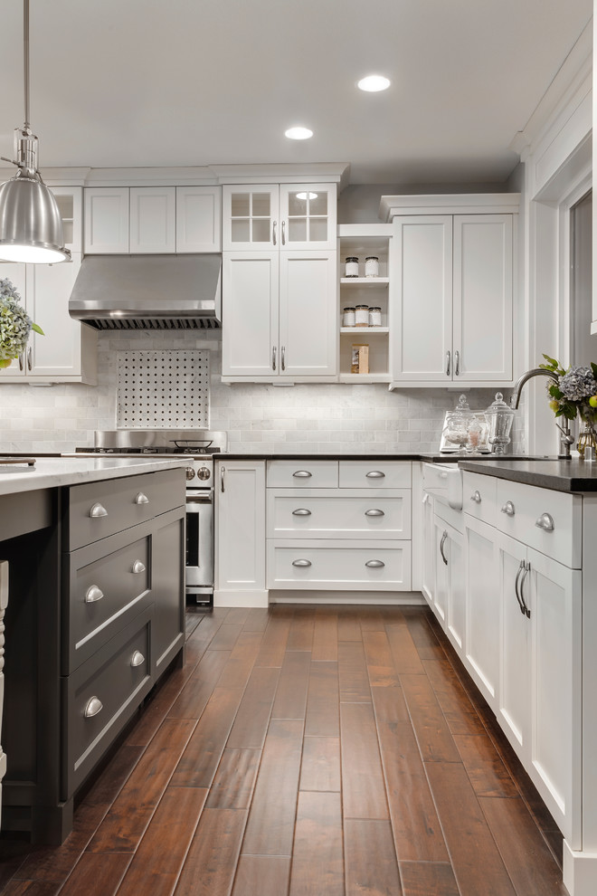 Inspiration for a large timeless l-shaped dark wood floor eat-in kitchen remodel in Denver with a farmhouse sink, shaker cabinets, white cabinets, solid surface countertops, gray backsplash, stone tile backsplash, stainless steel appliances and an island