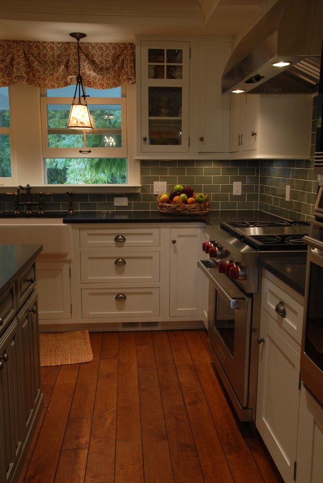 Inspiration for a medium sized classic u-shaped enclosed kitchen in Milwaukee with a belfast sink, shaker cabinets, white cabinets, green splashback, metro tiled splashback, stainless steel appliances, an island, medium hardwood flooring, brown floors and black worktops.