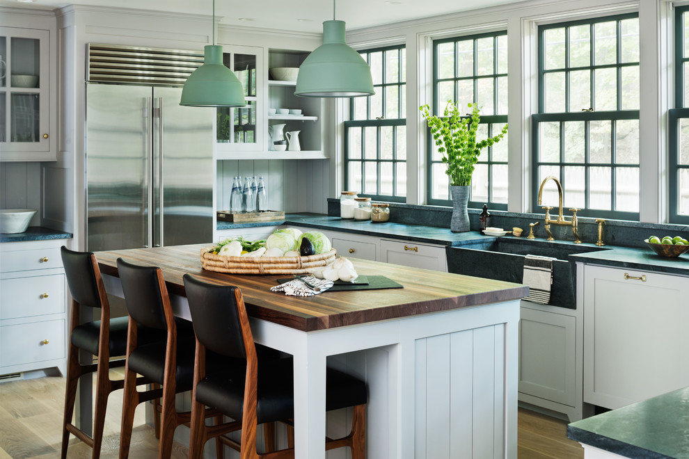 Klassische Küche in L-Form mit Landhausspüle, Glasfronten, weißen Schränken, Küchengeräten aus Edelstahl, hellem Holzboden und Kücheninsel in New York