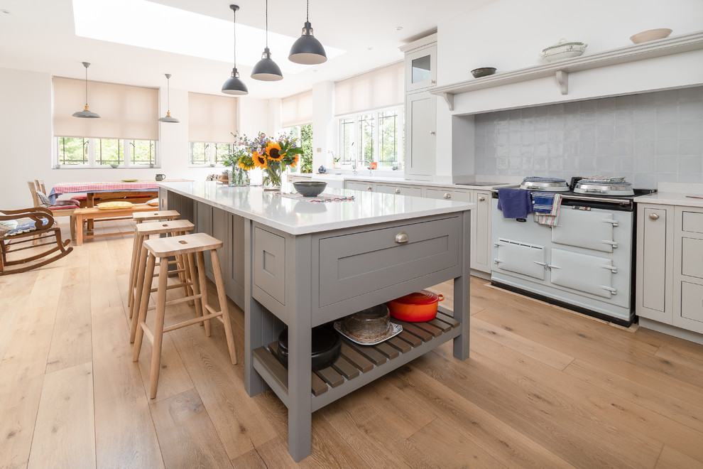 Photo of a large classic single-wall kitchen/diner in Buckinghamshire with a belfast sink, flat-panel cabinets, grey cabinets, engineered stone countertops, grey splashback, porcelain splashback, coloured appliances, light hardwood flooring, an island and white worktops.